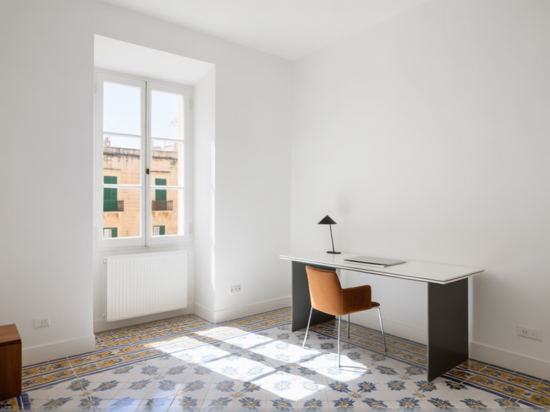 A clean, modern Maltese apartment interior, large windows, bright white walls, minimalist decor with a vibrant traditional Maltese tile floor. Sunlight streams in. A sleek desk setup is in the corner. Architectural Digest style, sharp focus on the contrast between old and new.